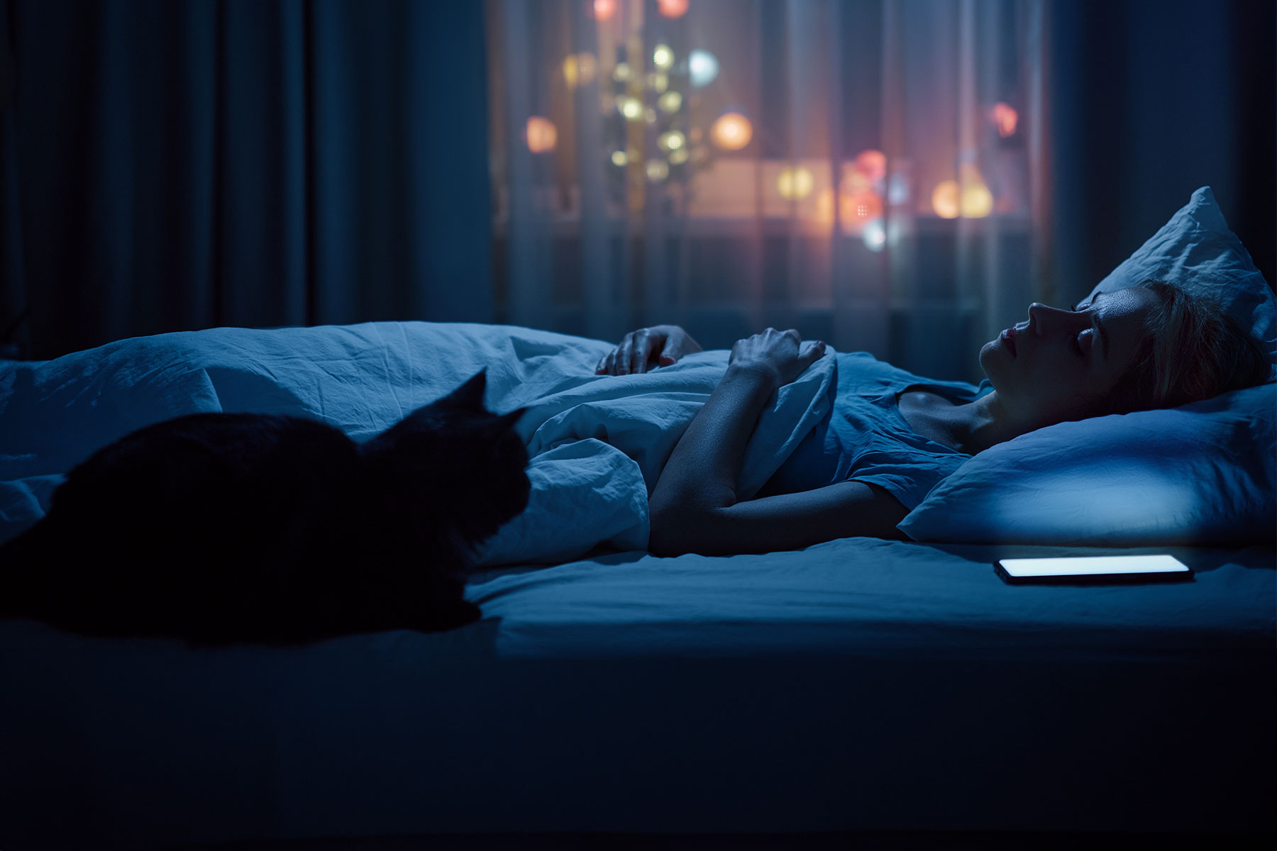 woman sleeping in dark bedroom, cat sitting at the foot of the bed