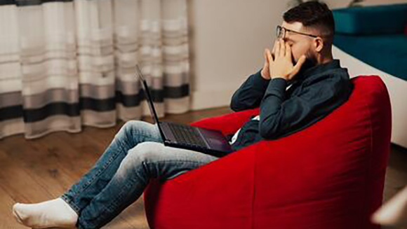 stressed male student sitting in a beanbag chair rubbing his eyes after starring at a laptop for hours