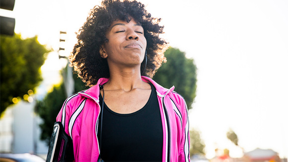 woman outdoors, possibly on a walk or jog, smiles up towards sky with eyes closed, possibly in relief