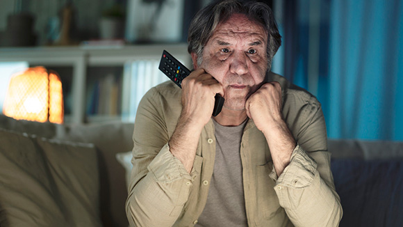 close up of an older man sitting on couch intently watching television, holding remote control
