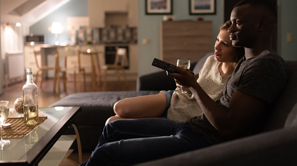 side view of a couple cuddled on couch watch television, woman holding a wine glass and leaning on man, man holding remote control