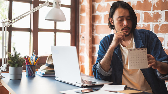 young man looking at a desk calendar, siting at desk in front of computer