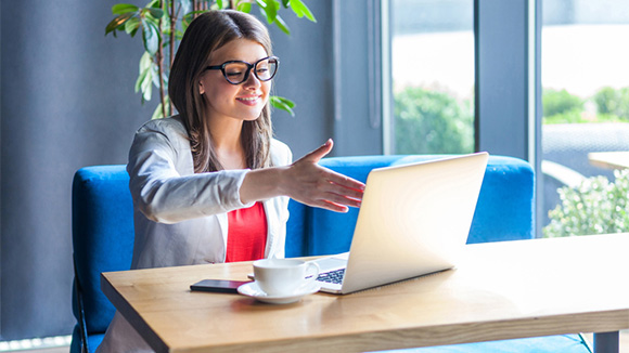 woman mimicking a handshake by holding out her hand to a laptop screen where she's on a virtual call