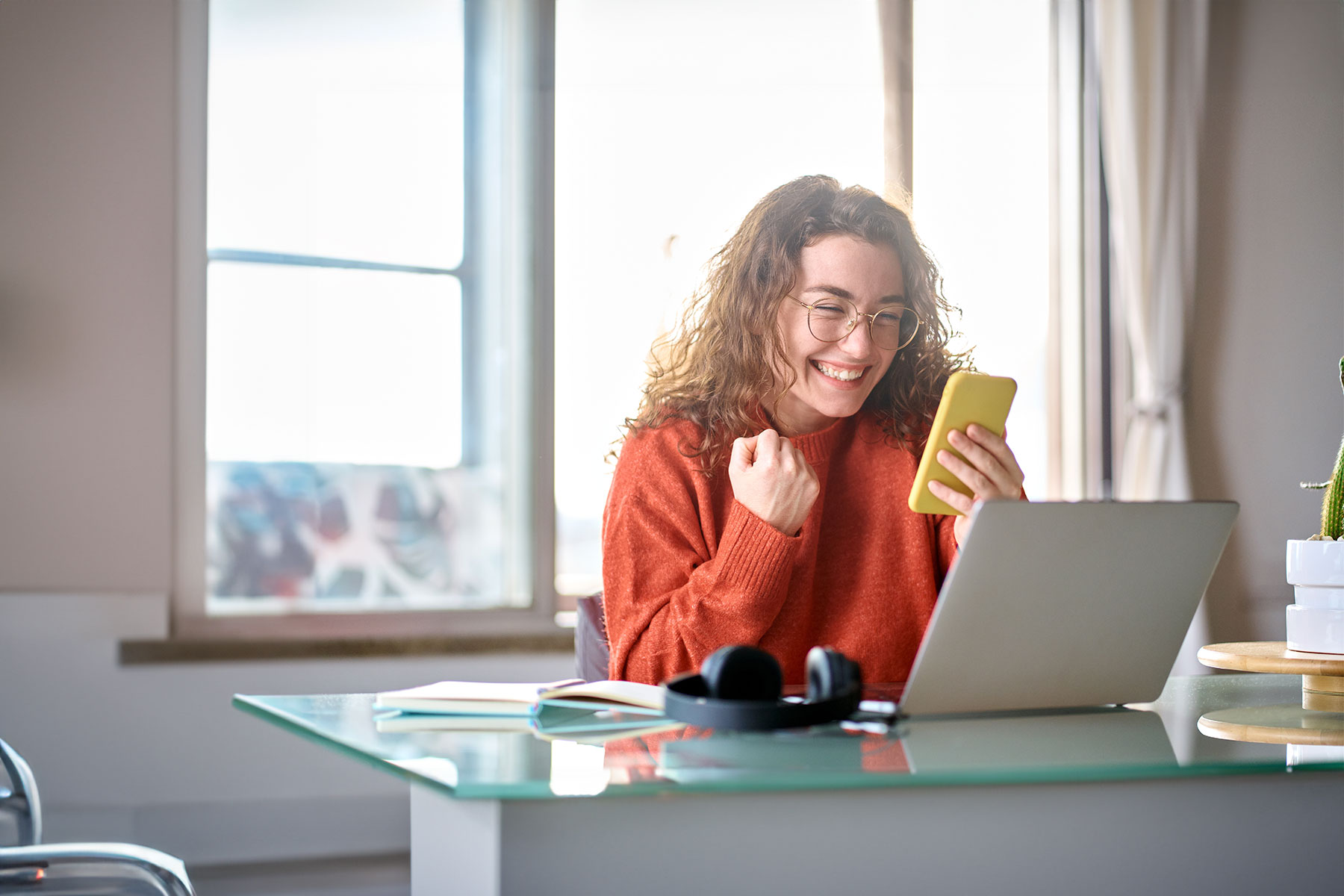 happy young woman sitting at desk in front of computer excited by what she's reading on her phone