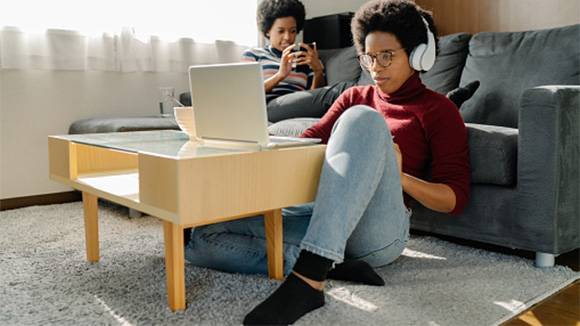 older sibling sits on the floor working on laptop as he leans against the couch, younger sibling is on phone while lounging on the couch