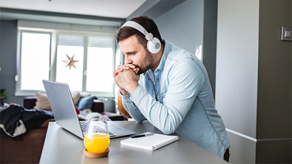young adult male leaning against kitchen counter wearing headphones, intensely watching video on laptop