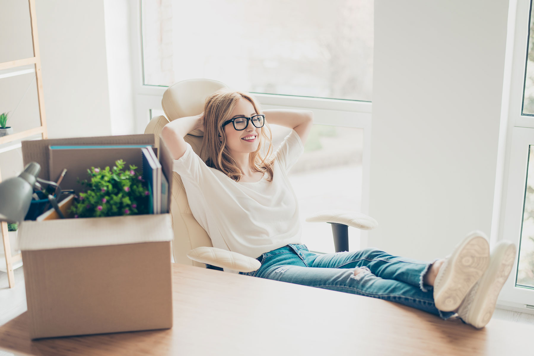 content woman lounges in office chair at desk in what appears to be a home office with small box of desk and office supplies sitting on desk