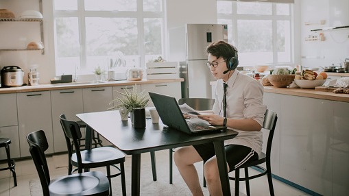 young man sitting at kitchen table on video call, wearing headset, professional work short, and lounge shorts