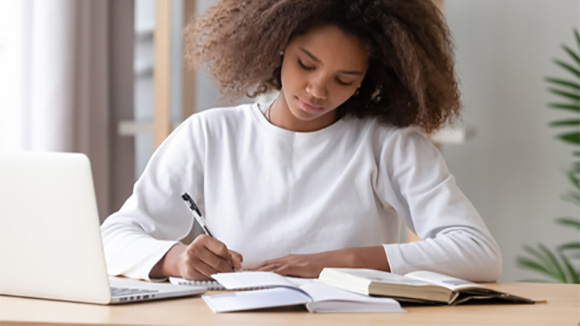 black female student writing notes, a laptop sitting in front of her