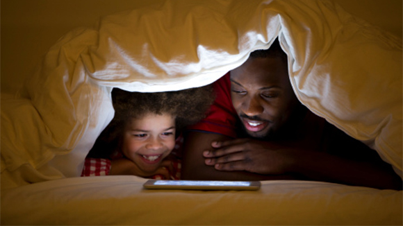 a black dad and his child in a blanket fort, watching a show on a tablet in front of them