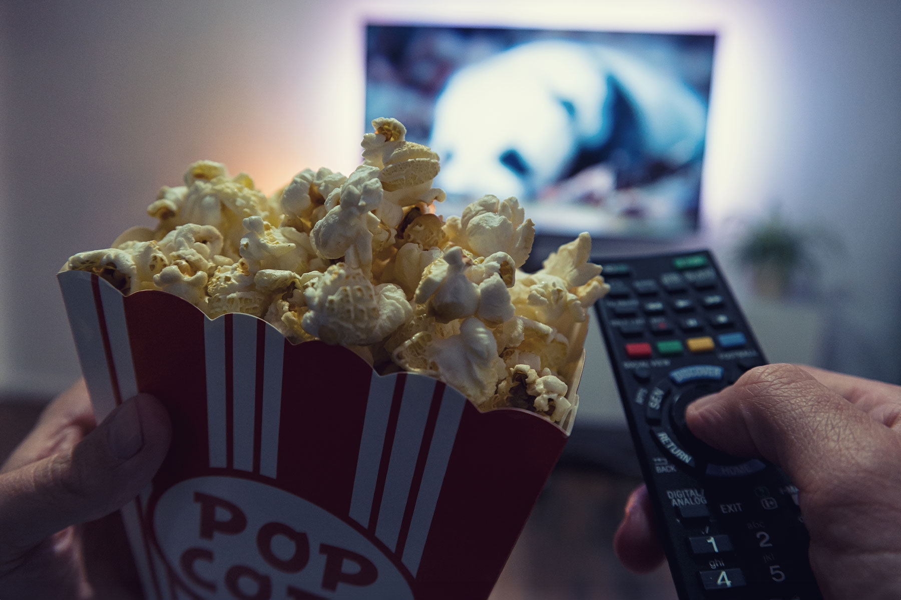close up of male hand holding television remote and container of popcorn