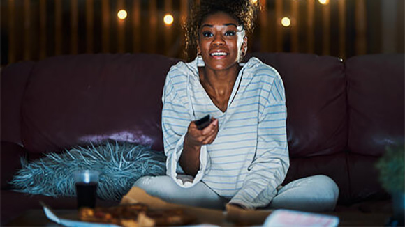 young woman sitting on the couch in the dark smiling and holding remote control, getting ready to enjoy some television or a movie