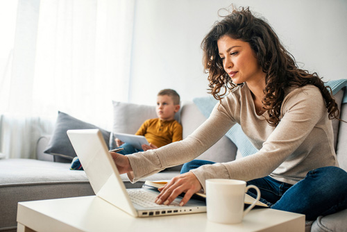 woman working on laptop while young boy in the background sits on couch playing on a tablet