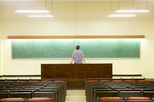 lone male teacher stands leaning against desk facing blackboard, empty classroom behind him