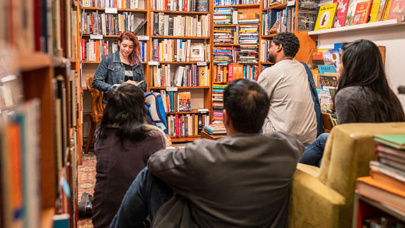 young woman standing in front of small crowd in used bookstore doing a reading