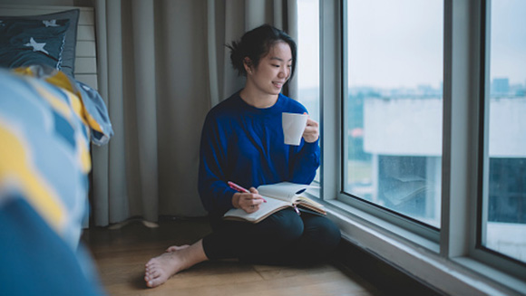 young woman sitting on floor of bedroom next to window with coffee and a journal