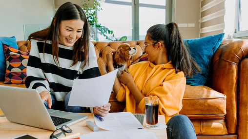 mother and daughter sitting on couch, mom working on laptop and reading papers while daughter plays with brown puppy