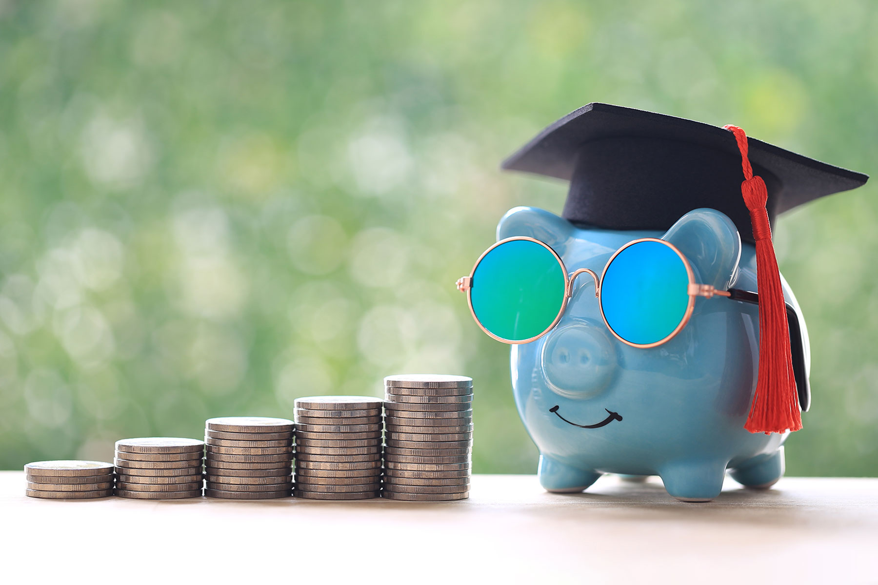 Graduation hat on piggy bank with stack of coins money on natural green background