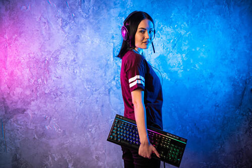 female gamer in front of a blue and purple background, looking over her shoulder, wearing headset and holding a multi-colored gaming keyboard
