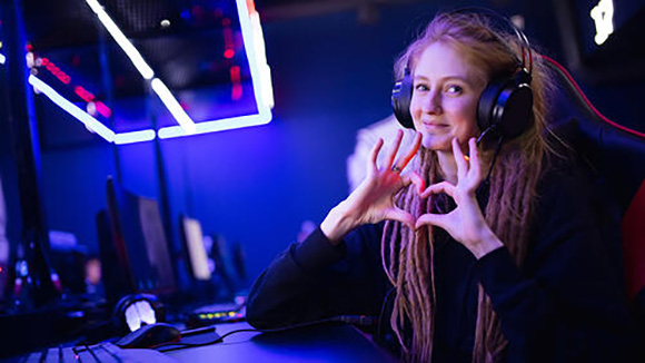 Female gamer wearing a headset sitting at a computer turns towards the camera and makes a heart sign out of her hands