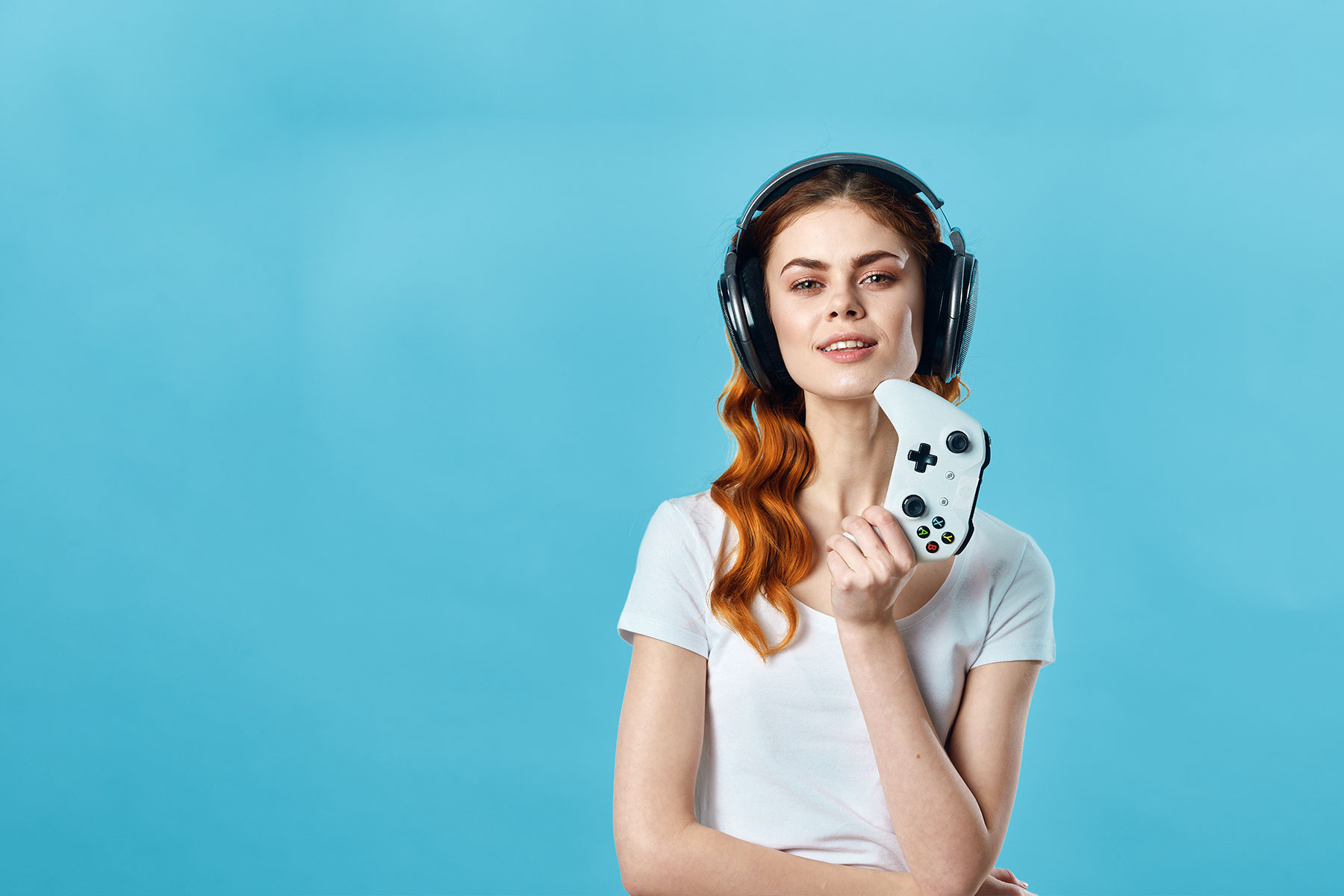 female gamer posing in front of a light blue background, wearing headphones and holding up a game controller