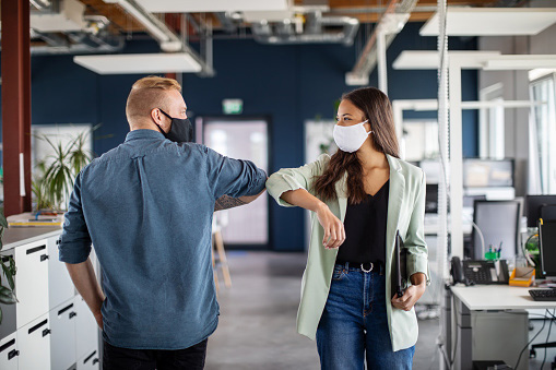 two co-workers passing each other in office hallway wearing masks and touching elbows