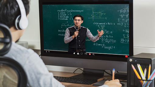 over the shoulder view of a young man watching a lecture on his computer, the instructor standing in front of a chalkboard with math equations on it