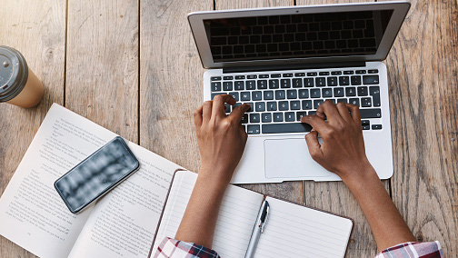 overhead view of someone using a laptop, open notebook and cellphone in front of them