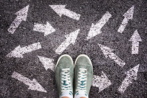 overhead closeup of a person's feet in tennis shoes stanfing on asphalt with multiple arrows pointing in a variety of directions