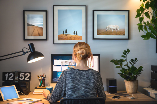 view of the back of a young blonde woman sitting at a desk on a computer, coffee cup in her left hand