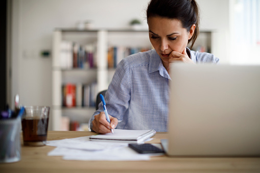 young brunette woman working pensively at a desk in front of a laptop, writing in a notebook