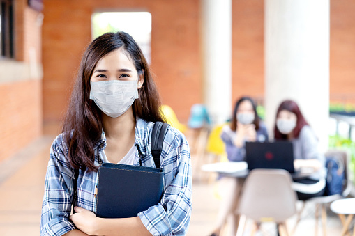 young female college student poses holding a book, wearing a mask during the covid-19 pandemic