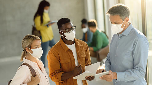 professor showing two students information on a piece of paper. they are in a busy lobby with everyone wearing masks