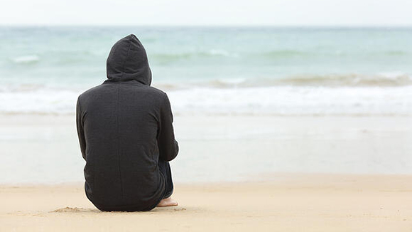 back view of a person sitting alone on beach wearing a dark sweatshirt and jeans, hood pulled up