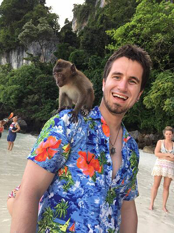 Professor David McDevitt smiles on a beach, wearing a tropical shirt while a small species of monkey sits on his shoulder