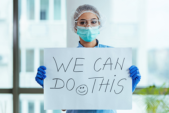 nurse wearing protective equipment including a mask, haircover, goggles, and gloves, holding up a sign reading "we can do this" with a drawn smiley face