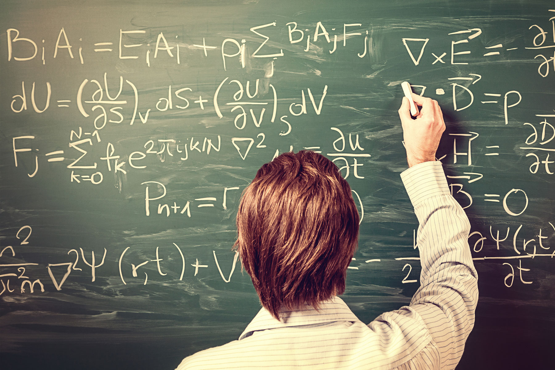 Rear view, close-up on young man standing back against green chalkboard working on complex equation