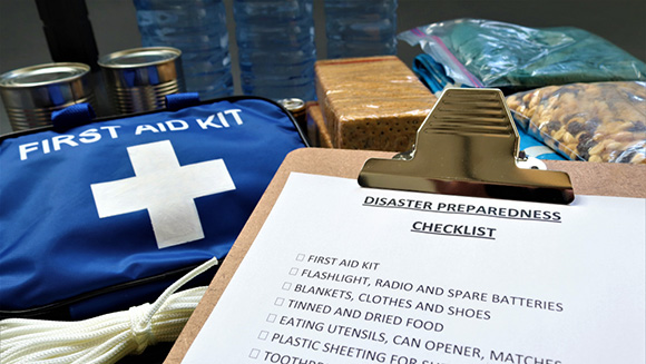 clipboard with emergency prepardness checklist in front of a variety of emergency kit supplies like bottled water, first aid kit, flashlights, and more