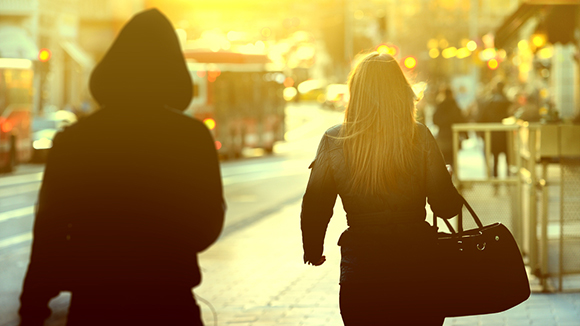 woman walking down busy city street being followed by a person in dark hoodie