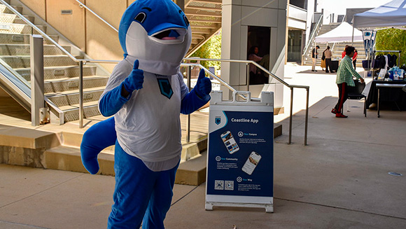 Coastline college dolphin mascot Fin stands at the Westminster campus next to a sign advertising the Coastline Mobile App