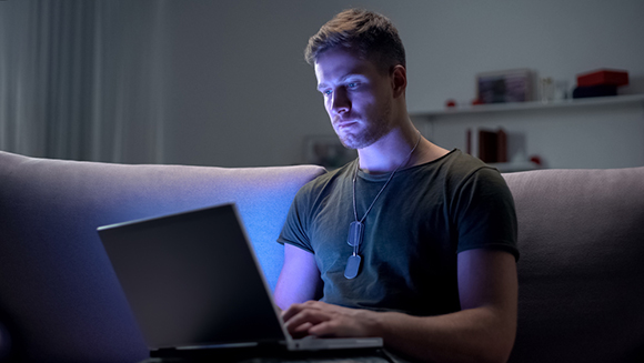 a service man sits on a couch in dim light looking at a laptop