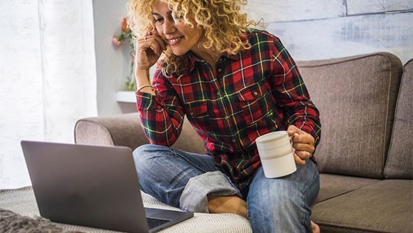 relaxed woman dressed cozily in a flannel and jeans sits on sofa with coffee looking at her laptop thats sitting on the coffee table in front of her