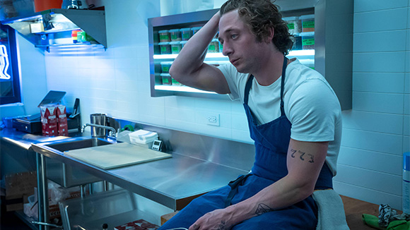 the bear - young man in restaurant kitchen, sitting on counter, hand in his hair, looking tired, stressed, and distraught