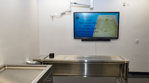 interior of an anatomy lap, stainless steel counter tops and dissection table with industrial lighting and a monitor on the wall reading "coastline college welcomes you to the john stauffer human anatomy suite"