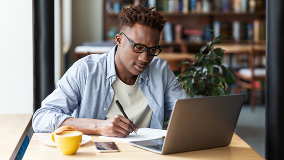 focused young man sitting at a cafe table working on laptop, taking notes down in a notebook