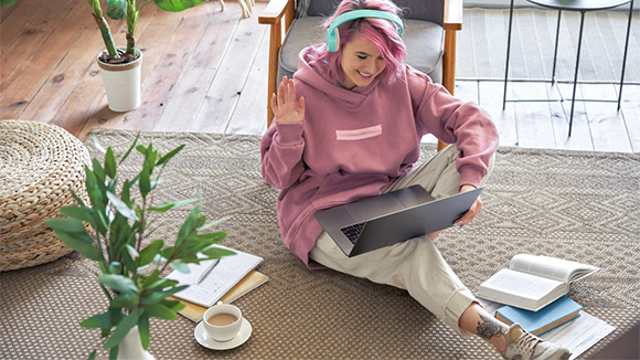 young pink-haired girl sitting on floor at home, on laptop, wearing headphones, smiling at screen as if on video call, notebooks, pen, coffee and other study supplies around her