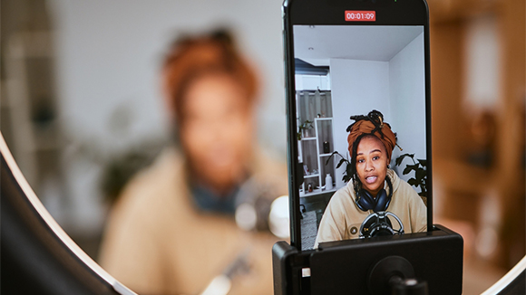 close up of phone set up in streaming set-up with ring light around it, showing a young, beautiful black woman making a video
