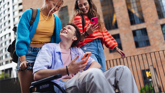 two young woman walking on street and laughing, one pushing a young man in wheelchair, also laughing and having fun