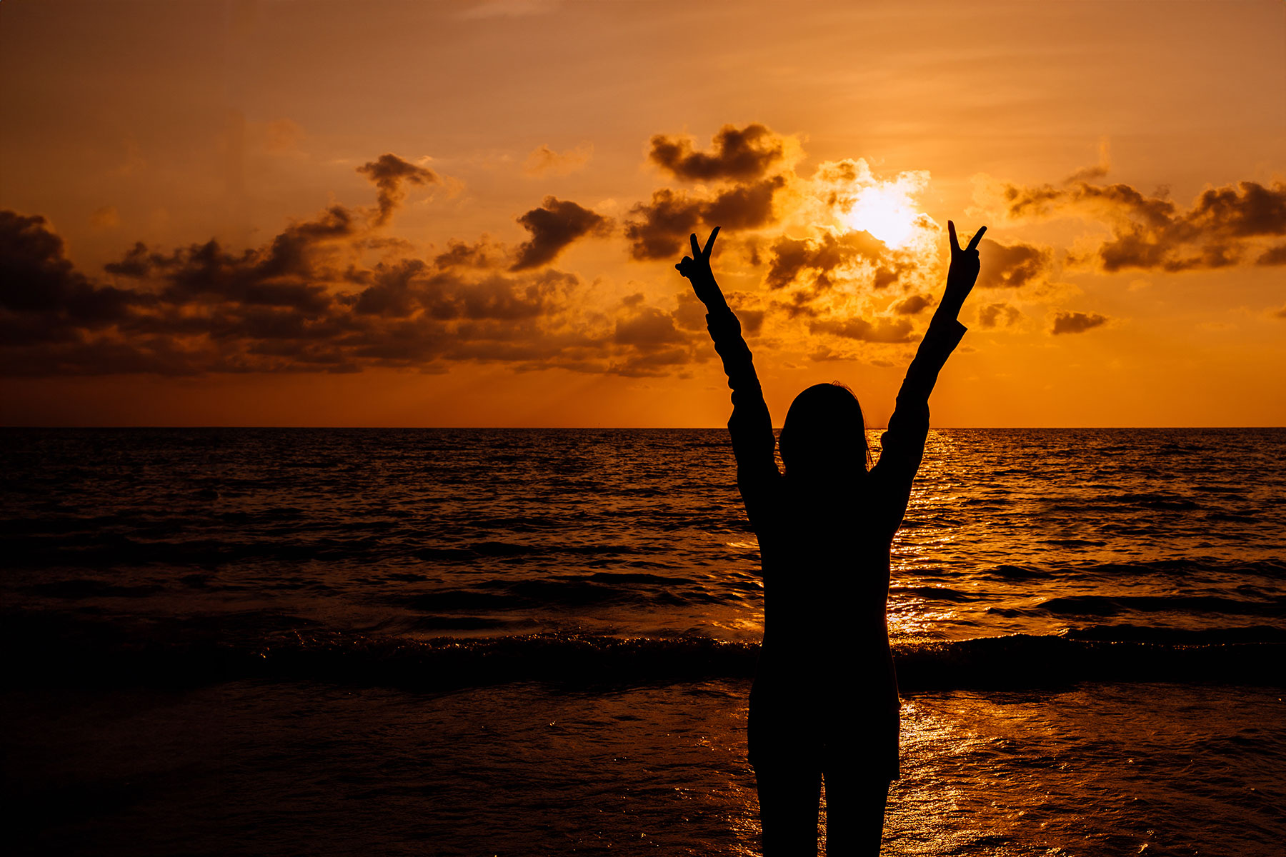 silhouette of an excited woman facing the sunset, holding hands up in peace signs