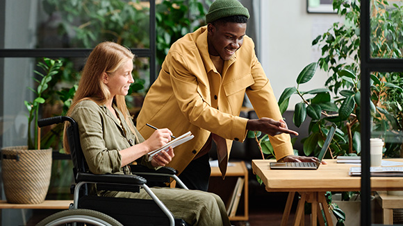 Female in wheelchair and young man standing next to her working on computer and taking notes in an open air office full of greenery and plants
